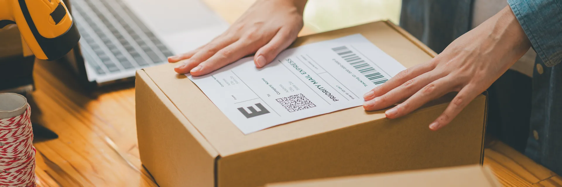 Hands smoothing an address label onto a cardboard box at a desk, beside a laptop and a barcode scanner.