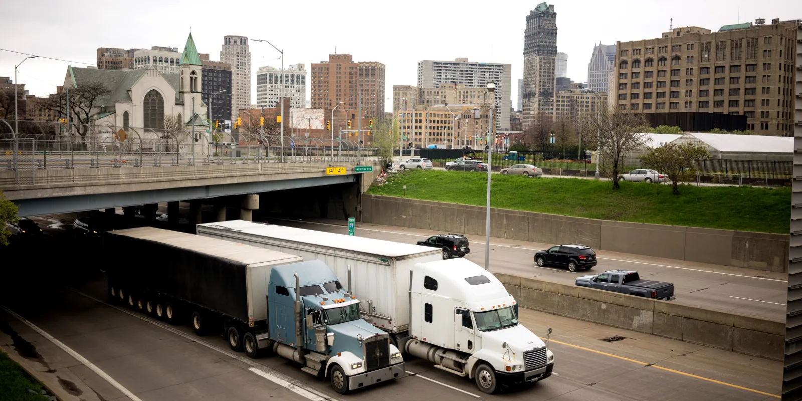 Two tractor-trailers on I-75 below an overpass, with the Detroit skyline behind.