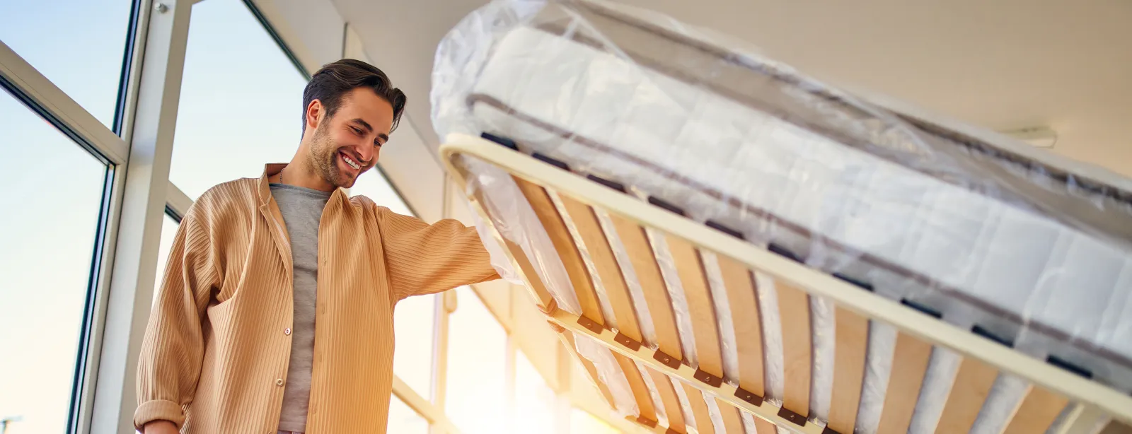 A man in a bed showroom lifting a slatted bed base, wrapped mattresses stacked around him.
